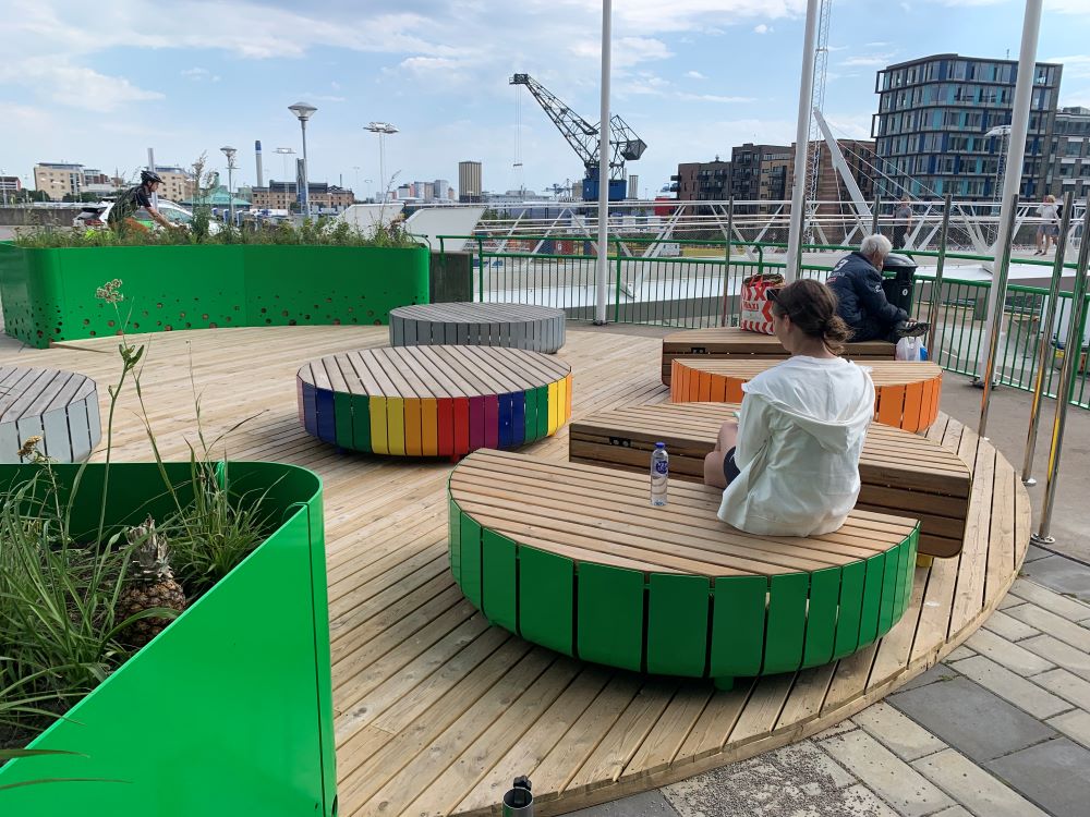 Personer sitter och slappnar av på färgglada, runda bänkar av trä på en utomhusplatå med modern stadsarkitektur i bakgrunden.
