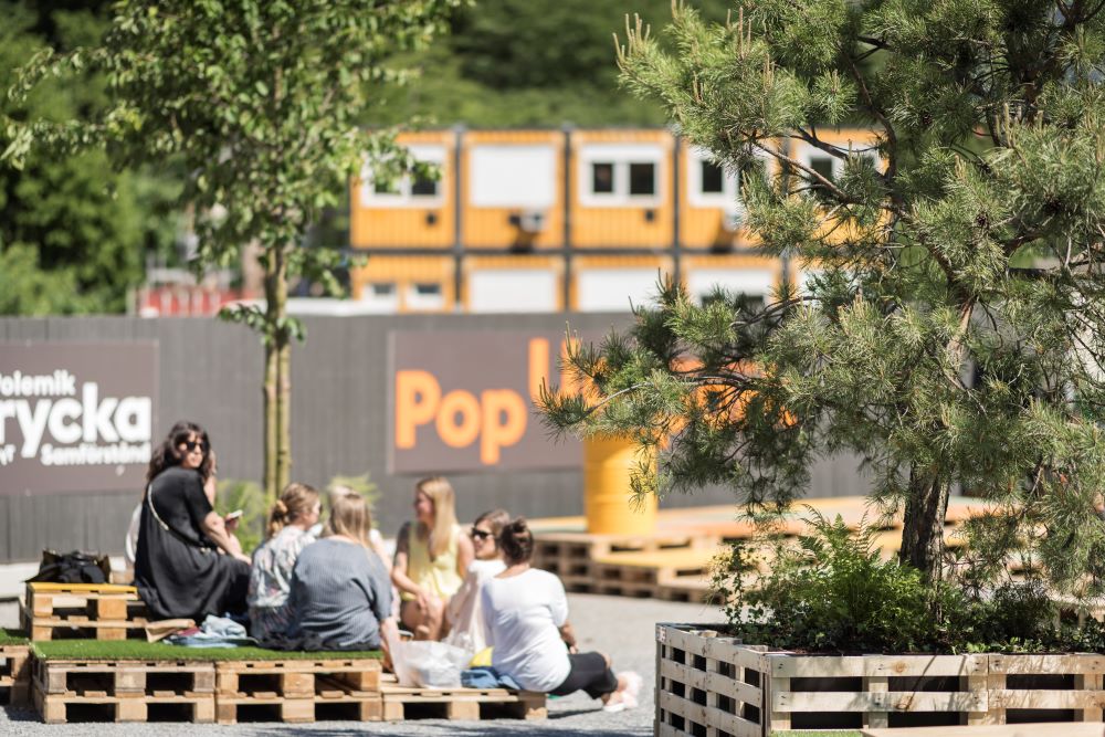 En grupp ungdomar sitter på träpallar i ett utomhusområde omgivet av träd, nära en skylt som säger "Pop Up".