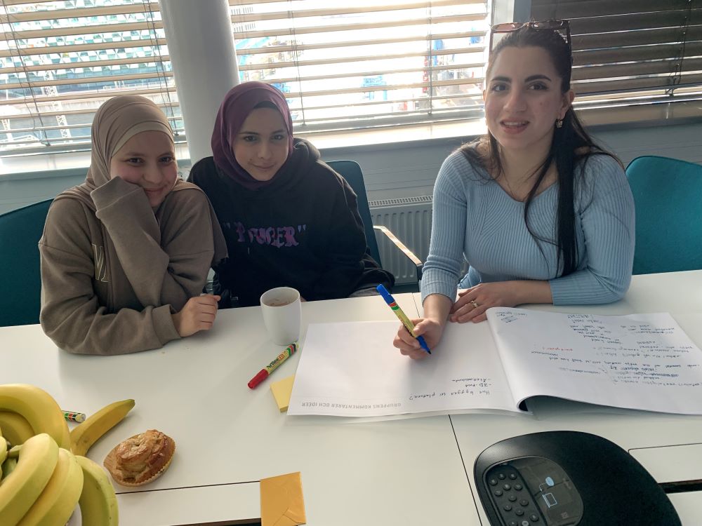 Tre ungdomar sitter vid ett bord och deltar i en workshop, med anteckningar på ett stort papper framför sig.