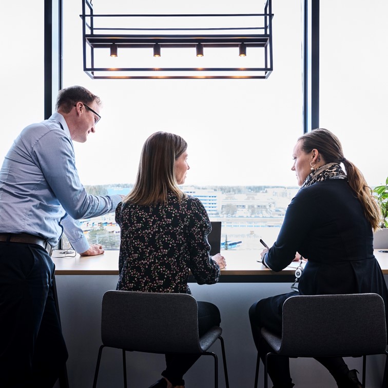 Tre personer som diskuterar i en öppen kontorsmiljö framför ett stort fönster med utsikt.