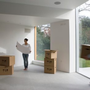 Omöblerad del av hus med flyttkartonger på golvet, en person som bär på en flyttkartong och en person som kollar på en ritning.