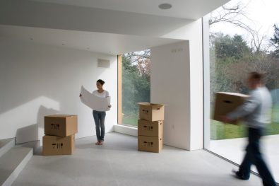 Omöblerad del av hus med flyttkartonger på golvet, en person som bär på en flyttkartong och en person som kollar på en ritning.