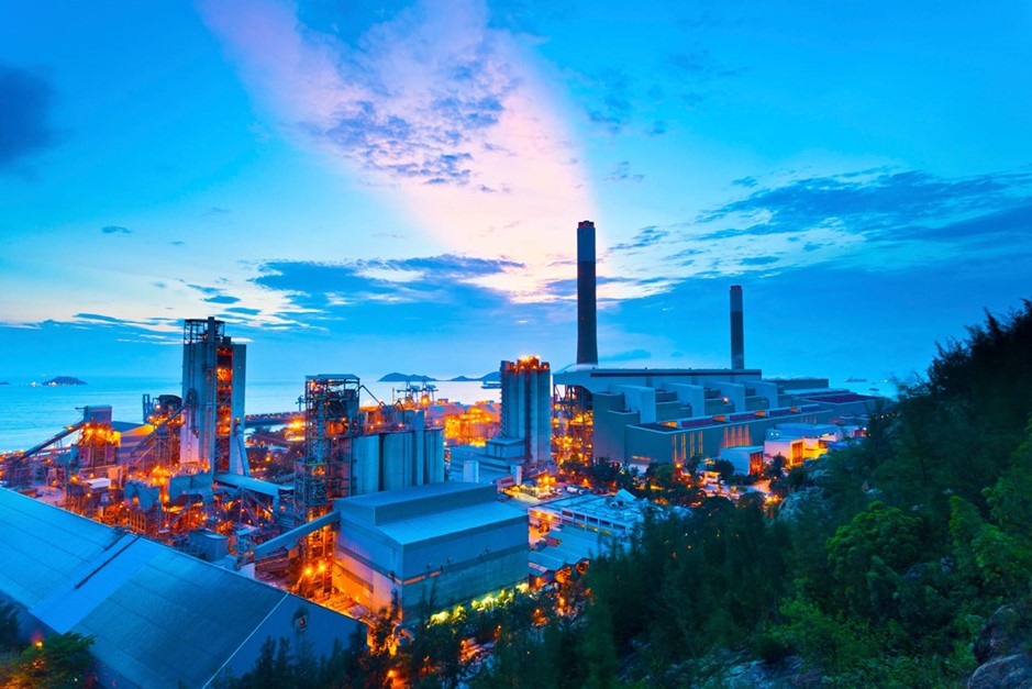 En stor industriell anläggning med flera byggnader och skorstenar är belägen vid havet under en blå kvällshimmel.