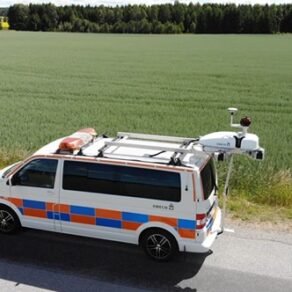 En Sweco-skåpbil med fastmonterad utrustning står parkerad vid en vägkant bredvid en stor åker på en solig dag.