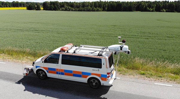 En Sweco-skåpbil med fastmonterad utrustning står parkerad vid en vägkant bredvid en stor åker på en solig dag.