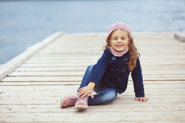 Ett barn med rosa mössa sitter på en brygga med vattnet i bakgrunden.