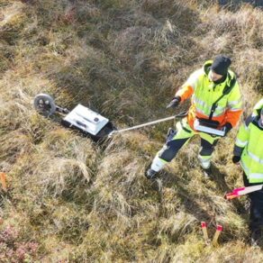 Två personer i varselkläder utför markundersökning med utrustning på ett gräsfält.