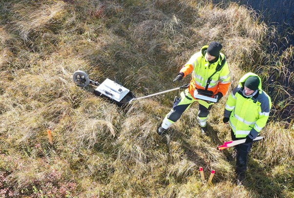 Två personer i varselkläder utför markundersökning med utrustning på ett gräsfält.