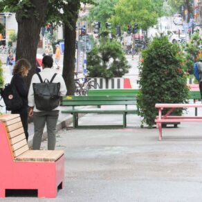 Stadsgata omvandlad till vistelseyta med färgglada bänkar och stora planteringar, några personer står, cyklar samt trafik syns i bakgrunden.