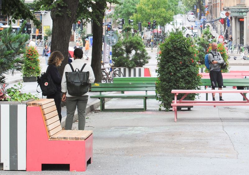 Stadsgata omvandlad till vistelseyta med färgglada bänkar och stora planteringar, några personer står, cyklar samt trafik syns i bakgrunden.