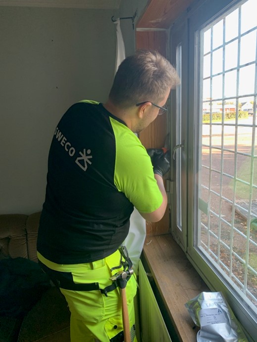 Person i varselkläder från Sweco arbetar vid ett fönster från insidan, vid ett gallerfönster med verktyg på fönsterbrädan.