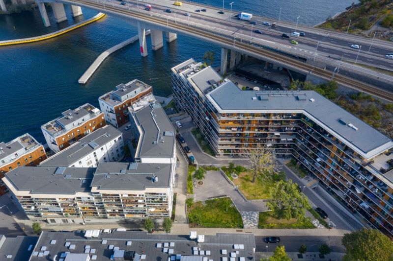 Flygfoto över moderna bostadshus på Bryggvägen, Gröndal i Stockholm, vid vattnet med innergård, intill en motorvägsbro och kaj.