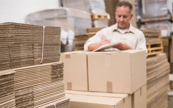 Staplar av kartonger och wellpapp i en lagermiljö, med en person som antecknar bakom lådorna.