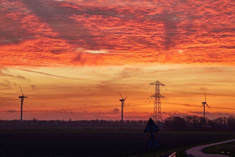 Vindkraftverk och kraftledning i landskap mot rödorange solnedgång.