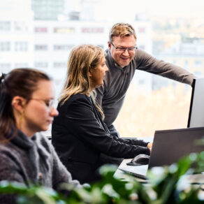 Tre kollegor samarbetar vid datorer på ett kontor, med fönster och stadsmiljö i bakgrunden.