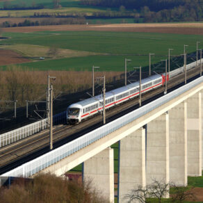 Höghastighetståg som kör på en elektrifierad järnvägsbro med höga betongpelare över ett öppet jordbrukslandskap.
