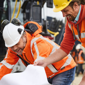 Två byggarbetare i skyddskläder granskar ritningar ovanpå stora stålrör på en aktiv byggarbetsplats.