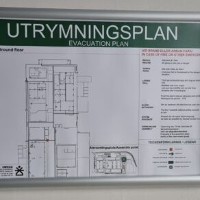 Utrymningsplan i aluminiumram, väggmonterad brandskylt med planritning över bottenvåning, markerade utrymningsvägar, samlingsplats samt instruktioner vid brand eller annan fara.