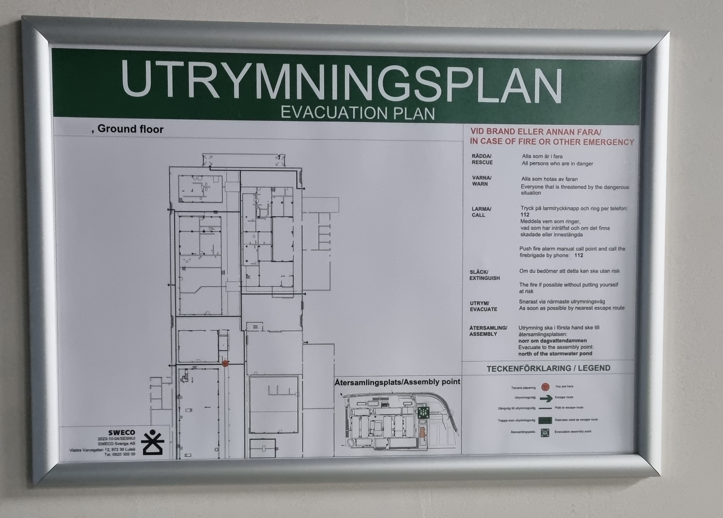 Utrymningsplan i aluminiumram, väggmonterad brandskylt med planritning över bottenvåning, markerade utrymningsvägar, samlingsplats samt instruktioner vid brand eller annan fara.