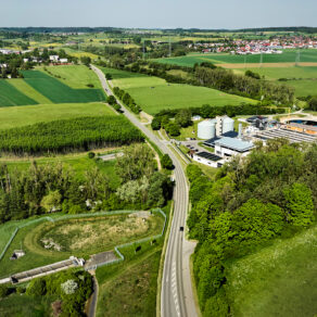 Flygbild över biogasanläggning i lantligt landskap med åkermark, skog och väg.