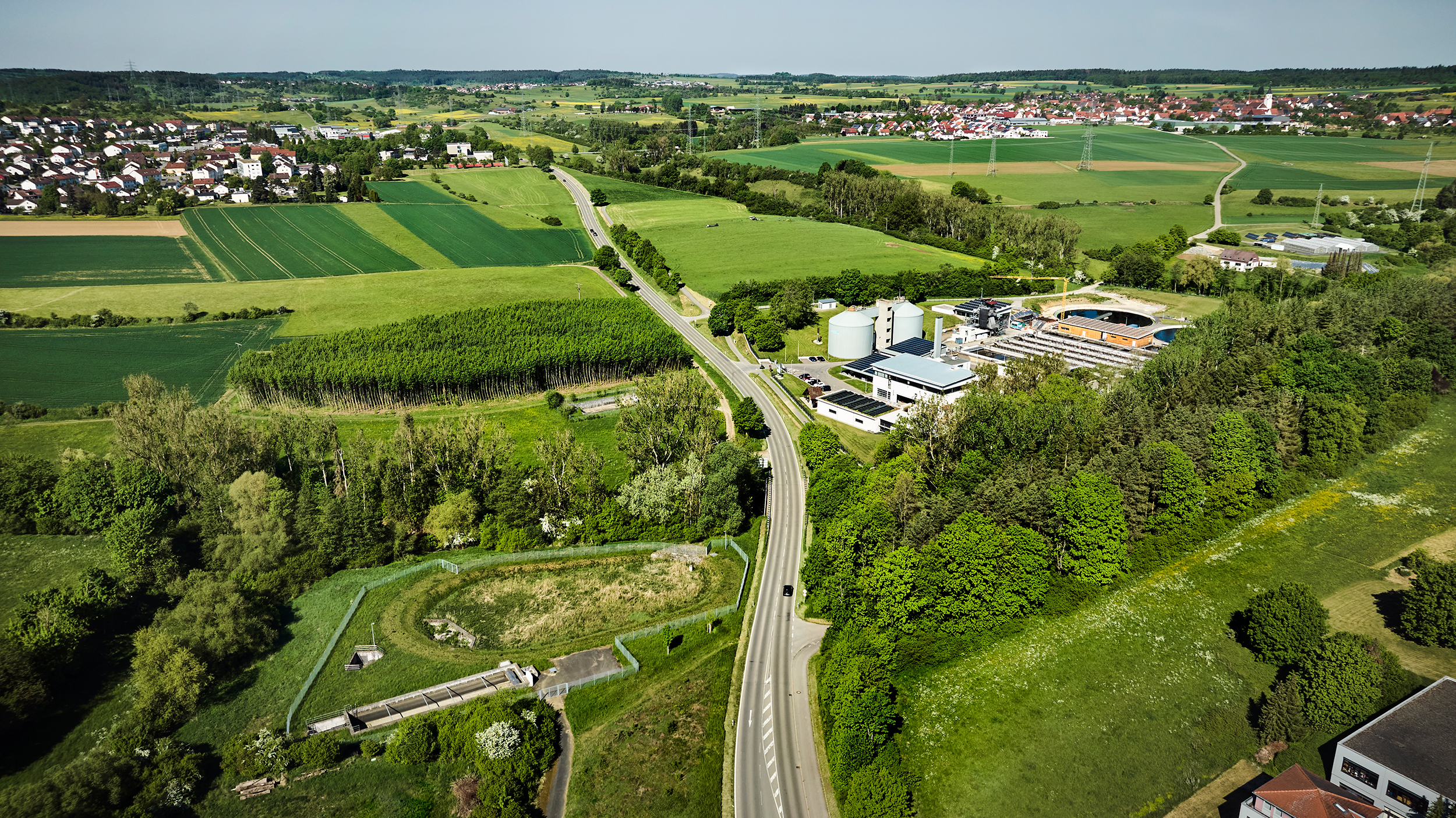 Flygbild över biogasanläggning i lantligt landskap med åkermark, skog och väg.