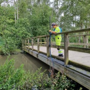 Miljötekniker tar vattenprov från träbro över å i grönskande park